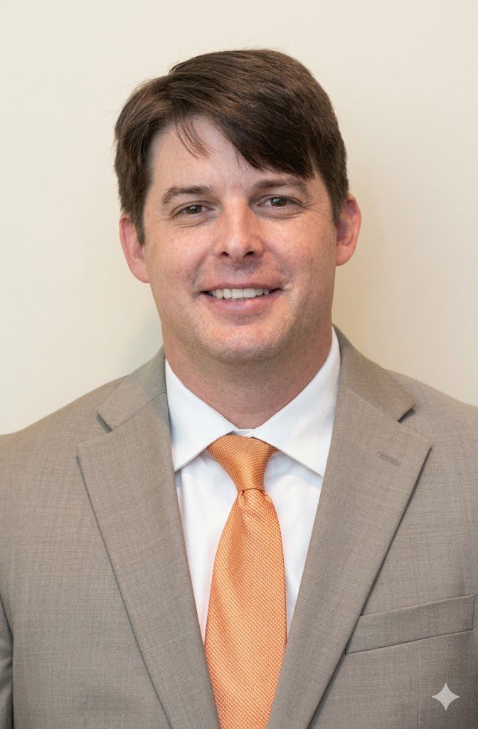 A smiling man in a light gray suit with an orange tie stands against a neutral background.