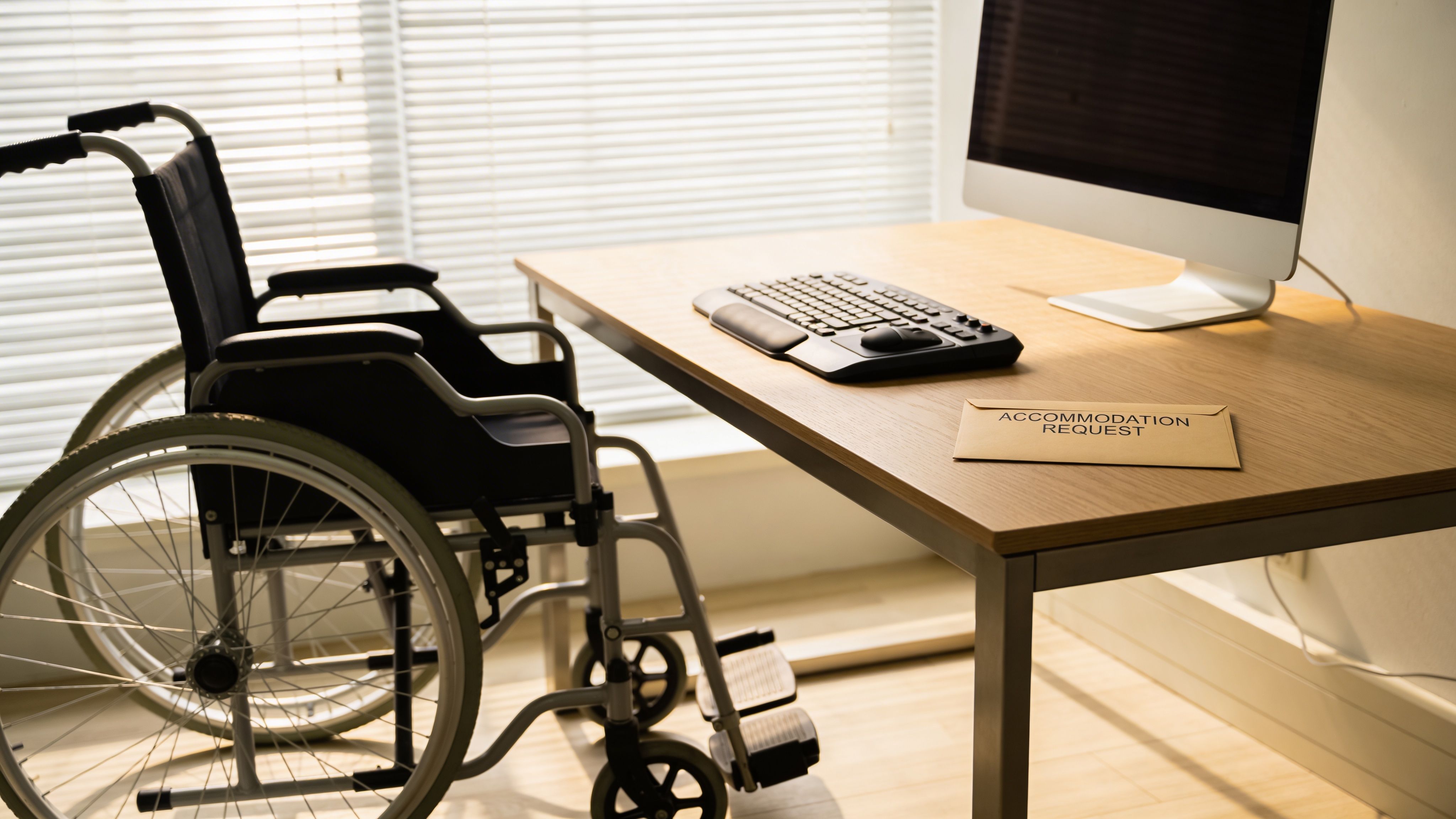 A wheelchair positioned next to an office desk with a computer and an accommodation request envelope.