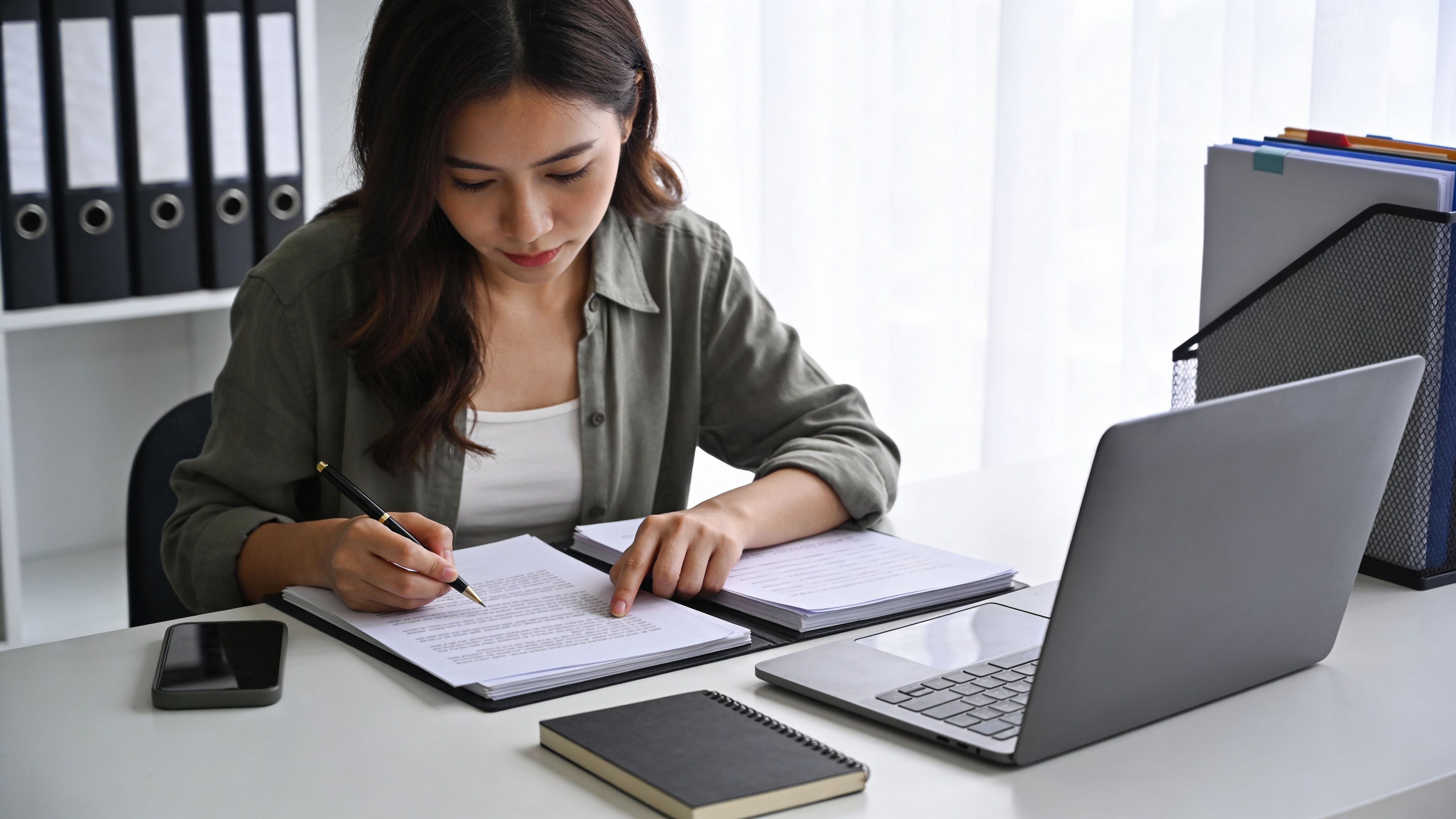 A professional woman meticulously reviewing and signing legal documents at her desk with a laptop and files.