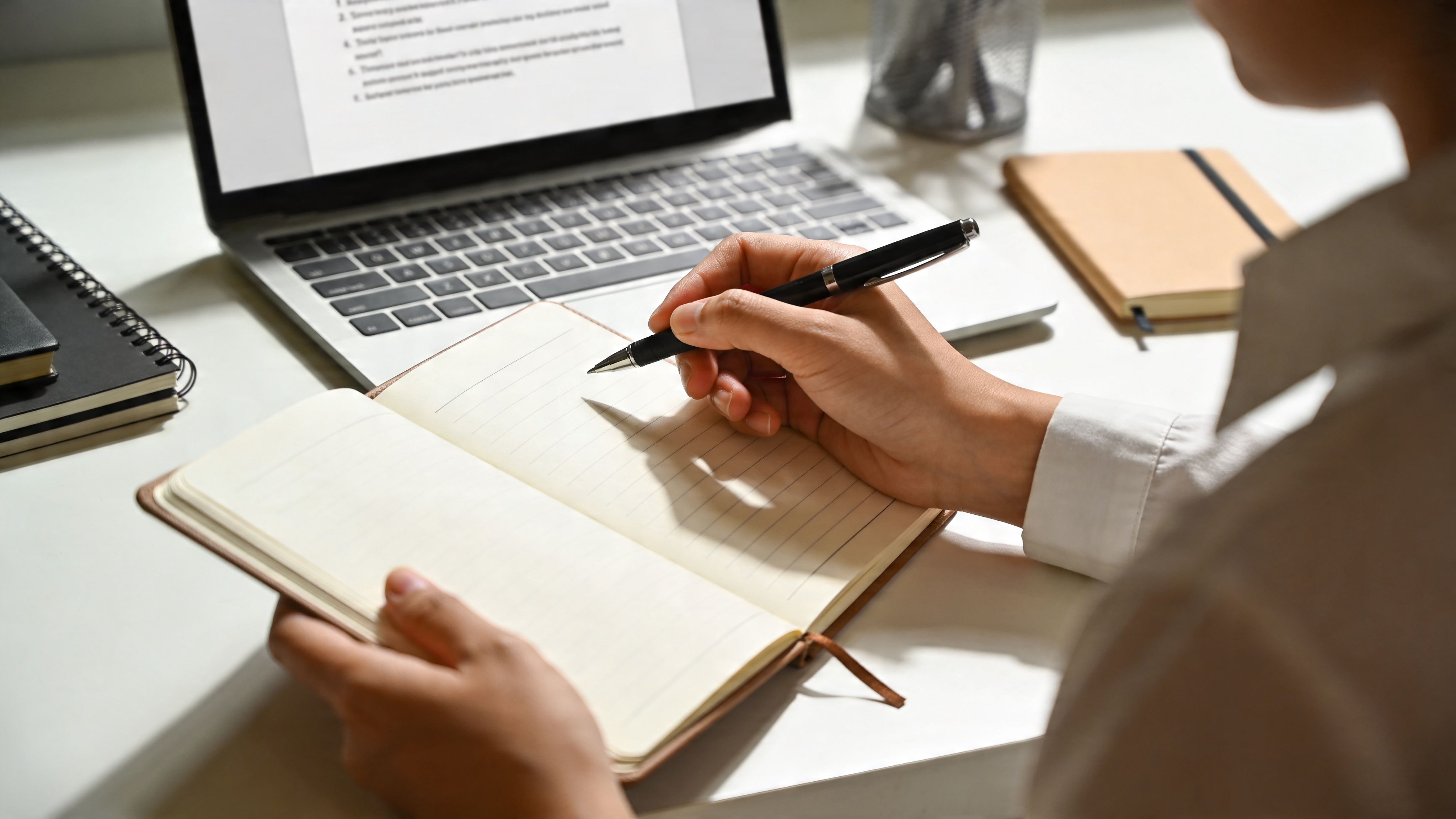 A professional writing in a notebook at a desk with a laptop and other stationery items.