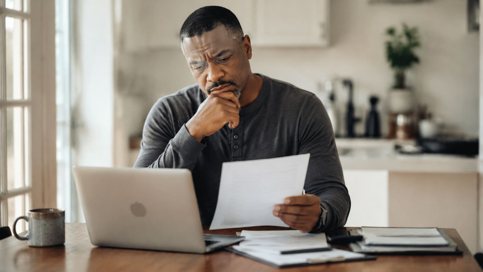A focused man sitting at a wooden table reviewing legal documents while working on his laptop computer.